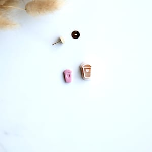 May include: Two pairs of earrings featuring a coffee cup design. One pair is pink and the other is white.