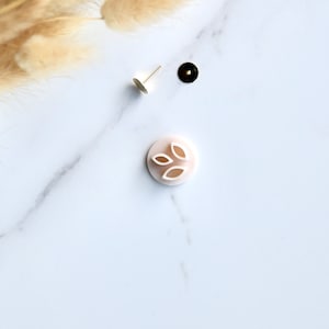 May include: A white plastic leaf-shaped cookie cutter with three leaves. The cutter is on a white marble surface with a gold pin and a black button.