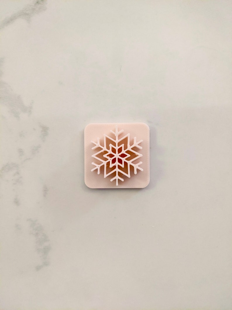 K&ouml;nnte beinhalten: Eine rosa Silikonform mit einem Schneeflockenmuster. Die Schneeflocke ist wei&szlig; umrandet und hat eine rote Mitte.