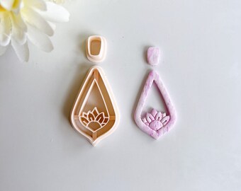 May include: Two cookie cutters in the shape of a teardrop with a floral design in the center. The cutter on the left is white and the cutter on the right is pink.