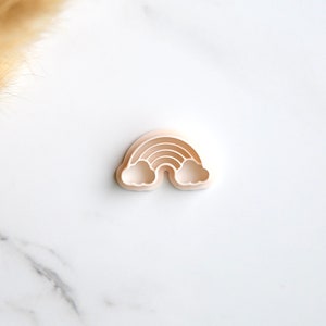 Puede incluir: Un cortador de galletas de plástico rosa claro en forma de arcoíris con dos nubes a cada lado del arcoíris.