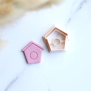 May include: A pink plastic cookie cutter shaped like a birdhouse with a round hole in the front. The cutter is on a white surface.