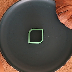 May include: A green leaf-shaped cookie cutter on a black plate. The plate is on a brown cork surface.