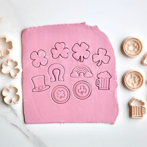 May include: A set of 14 cookie cutters featuring various St. Patrick's Day themes, including shamrocks, rainbows, beer mugs, hats, and horseshoes. The cutters are arranged on a pink fondant sheet and a white surface.