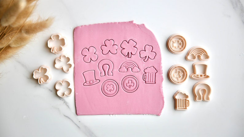 May include: A set of 14 cookie cutters featuring various St. Patrick's Day themes, including shamrocks, rainbows, beer mugs, hats, and horseshoes. The cutters are arranged on a pink fondant sheet and a white surface.