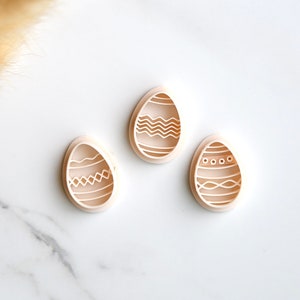 May include: Three white plastic Easter egg cookie cutters with different patterned designs. The cutters are on a white marble surface.