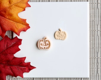 May include: Two pumpkin-shaped cookie cutters, one with a carved-out face and the other a solid pumpkin shape. The cutters are light brown and sit on a white surface, with fall leaves in the background.