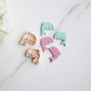 May include: Set of five dolphin cookie cutters, two are white and three are colored. The colored cutters are light blue, pink, and pink.