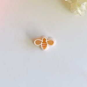 May include: A white plastic cookie cutter shaped like a bee with orange stripes.