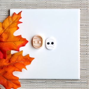 May include: Two Halloween-themed cookie cutters, one shaped like a mummy with black eyes and the other shaped like a mummy's face with two eyes.
