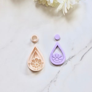 Könnte beinhalten: Zwei tropfenförmige Ausstecher, einer weiß und einer lila, mit einem Blumendesign in der Mitte.