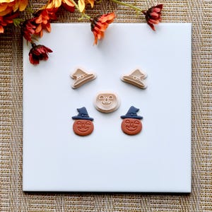May include: Five Halloween-themed cookie cutters on a white surface. Two are pumpkin-shaped with a witch's hat on top, and two are just pumpkins. The fifth is a plain pumpkin shape.