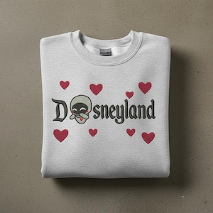 May include: A light gray sweatshirt with the word "Dsneyland" embroidered in black, featuring a cartoon skull character and red hearts. The sweatshirt is folded neatly on a neutral surface.