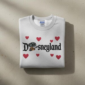 May include: A light gray sweatshirt with the word "Disneyland" in black script, featuring a cartoon character and red heart accents. The sweatshirt is folded neatly on a neutral surface.