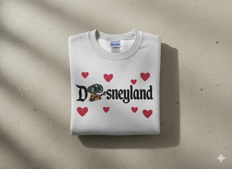 May include: A light gray sweatshirt with the word "Disneyland" in black script, featuring a cartoon character and red heart accents. The sweatshirt is folded neatly on a neutral surface.