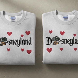 May include: Two folded light gray sweatshirts with the word "Disneyland" in black script, featuring a cartoon character and red hearts. The sweatshirts have a crew neck and are folded neatly.