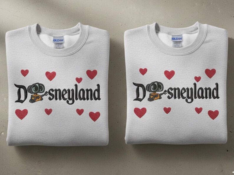 May include: Two folded light gray sweatshirts with the word "Disneyland" in black script, featuring a cartoon character and red hearts. The sweatshirts have a crew neck and are folded neatly.