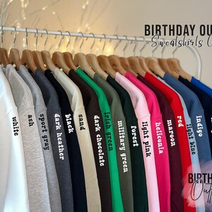 May include: A rack of sweatshirts in various colors, including white, ash, sport gray, black, sand, dark chocolate, Irish green, and more. Each sweatshirt is labeled with its color name in a simple font. The text "BIRTHDAY OUTFITS" and "Sweatshirts Colors" is visible.