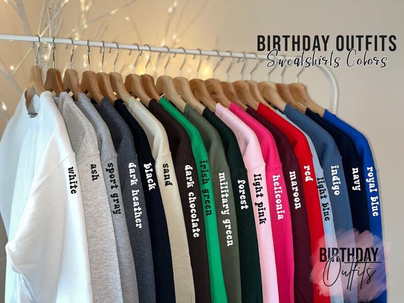 May include: A rack of sweatshirts in various colors, including white, ash, sport gray, black, sand, dark chocolate, Irish green, and more. Each sweatshirt is labeled with its color name in a simple font. The text "BIRTHDAY OUTFITS" and "Sweatshirts Colors" is visible.