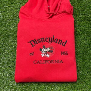 May include: A red hooded sweatshirt with the word "Disneyland" embroidered in black script. Below the word is a graphic of Mickey and Minnie Mouse, with the text "est 1955 CALIFORNIA". The sweatshirt is folded on a green surface.