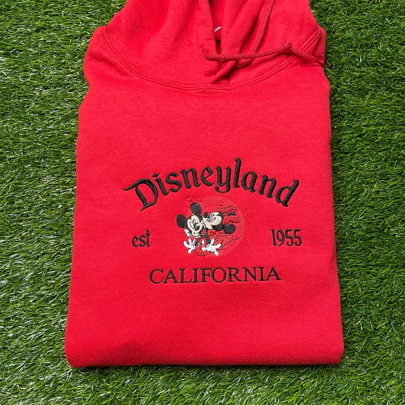 May include: A red hooded sweatshirt with the word "Disneyland" embroidered in black script. Below the word is a graphic of Mickey and Minnie Mouse, with the text "est 1955 CALIFORNIA". The sweatshirt is folded on a green surface.