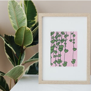 May include: A framed print of a pink background with green heart-shaped leaves hanging down. The leaves are outlined in black and have a speckled texture.