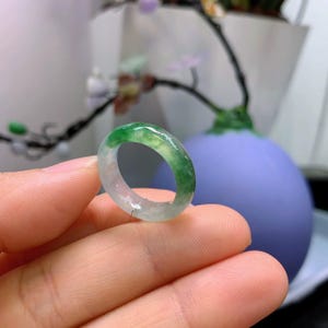 May include: A translucent jade ring with a gradient of green and white hues. The ring is held in a hand, showcasing its polished surface and the natural variations in color. The ring is a piece of jewelry.