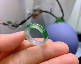 May include: A translucent jade ring with a vibrant green and white color gradient. The ring is held in a hand, showcasing its polished surface and the natural variations in the stone. The ring's design is simple and elegant.