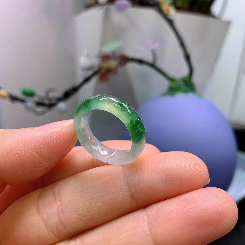 May include: A translucent jade ring with a vibrant green and white color gradient. The ring is held in a hand, showcasing its polished surface and the natural variations in the stone. The ring's design is simple and elegant.
