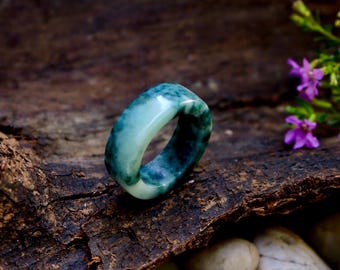 May include: A jade ring with a marbled appearance, showcasing shades of green and white. The ring is set against a dark, textured background, with small purple flowers and white stones in the background. The ring is a band style.