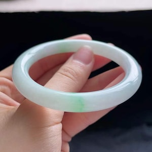 Op de afbeelding: Een jade armband, met een glad, gepolijst oppervlak met een doorschijnende, lichtgroene en witte kleur. De cirkelvormige armband wordt in een hand gehouden, wat het elegante ontwerp en de natuurlijke steensamenstelling benadrukt.