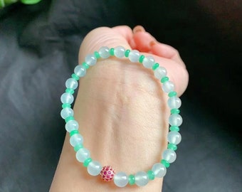 May include: A beaded bracelet featuring light blue and green beads. The bracelet has a central bead with a cluster of small red stones. The bracelet is worn on a wrist.