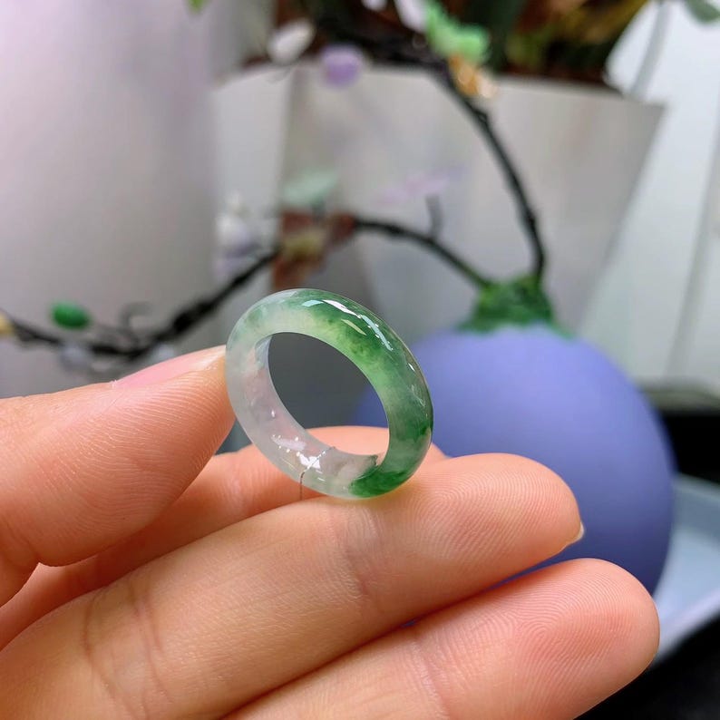 May include: A translucent jade ring with a mix of green and white hues. The ring is smooth and polished, showcasing the natural variations in the stone. The ring is held in a hand, with a blurred background.