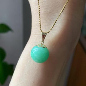 May include: A gold-coloured necklace featuring a light green, spherical pendant. The pendant has a decorative gold-coloured cap. The chain is a delicate ball chain, creating a simple and elegant piece of jewellery.