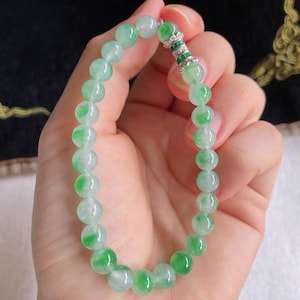 May include: A jade bracelet featuring round, translucent beads in shades of green and white. The bracelet is held in a hand, showcasing the polished stones and the silver clasp with green and clear accents. The overall aesthetic is elegant and natural.