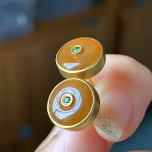 Op de afbeelding: Een paar gouden oorbellen met oranje jade stenen en groene edelstenen die in het midden van elke steen zijn gezet.