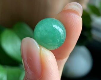 Könnte beinhalten: Ein runder, polierter Edelstein aus jadegrünem Jade. Der Edelstein wird zwischen zwei Fingern gehalten, wodurch seine glatte, reflektierende Oberfläche hervorgehoben wird. Die Farbe ist ein lebendiges, durchscheinendes Grün.