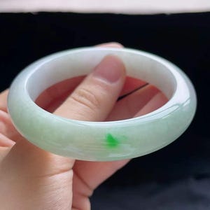 Op de afbeelding: Een jade armband in lichtgroen en wit. Het gladde, gepolijste oppervlak van de armband wordt geaccentueerd door een kleine, levendige groene vlek. De armband wordt in een hand gehouden, wat het cirkelvormige ontwerp laat zien. Dit stuk is een soort sieraad.