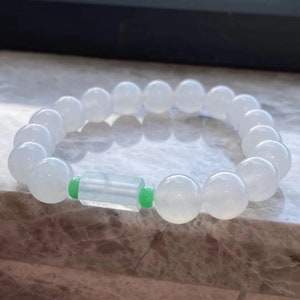 May include: A white jade bracelet with a green jade bead accent. The bracelet is made of round beads and is approximately 7 inches in length.