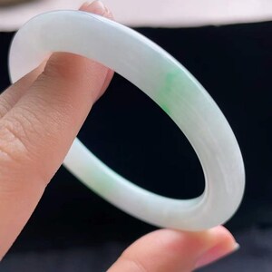 Op de afbeelding: Een gladde, witte jade armband met een vleugje groen. De cirkelvormige armband wordt omhoog gehouden en toont zijn gepolijste oppervlak en subtiele kleurvariaties. Een elegant sieraad.
