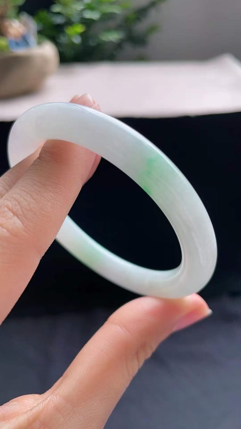 Op de afbeelding: Een gladde, witte jade armband met een vleugje groen. De cirkelvormige armband wordt omhoog gehouden en toont zijn gepolijste oppervlak en subtiele kleurvariaties. Een elegant sieraad.