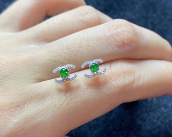 May include: A pair of silver earrings with a green gemstone in the center of each earring. The earrings are shaped like the letter 'C' and are decorated with small white gemstones.