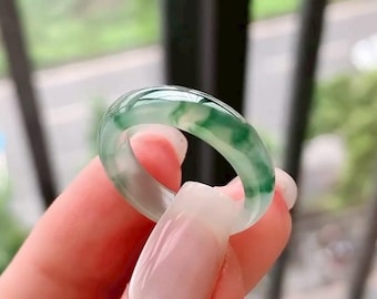 May include: A translucent jade ring with a smooth, polished surface. The ring features a blend of light green and white hues, creating a marbled effect. The ring is held up by a person's fingers.
