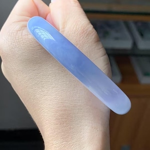 May include: A smooth, lavender-colored jadeite bangle bracelet. The polished surface reflects light, showcasing the natural variations in the stone. The bracelet is held in a hand, highlighting its shape and color.