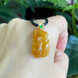 May include: A gold-colored pendant necklace featuring a carved figure. The pendant is suspended from a black cord and has a gold-colored clasp with a small green stone. The figure is a warm yellow color.