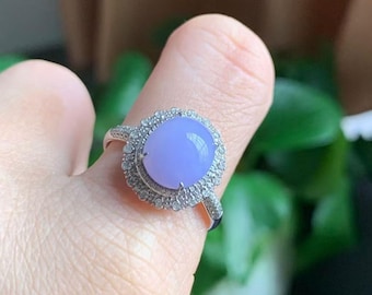 May include: A silver ring featuring a large, oval-shaped, lavender-colored gemstone. The gemstone is surrounded by a halo of smaller, clear stones. The ring is displayed on a finger against a blurred background of green foliage.