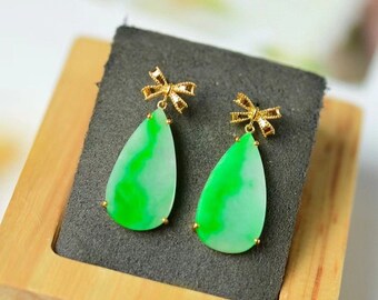 May include: A pair of jade drop earrings with gold bow accents. The earrings are teardrop shaped and feature a green and white jade stone.