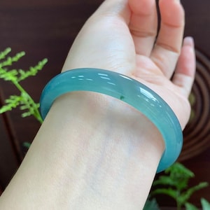 Op de afbeelding: Een doorschijnende, lichtblauwe jadeiet armband. Het gladde, gepolijste oppervlak reflecteert het licht en toont de natuurlijke kleurvariaties. Een klassiek sieraad.