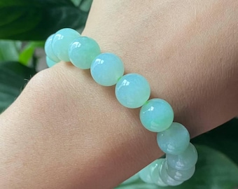 Könnte beinhalten: Ein Armband aus hellgrünen Jadeperlen. Das Armband besteht aus großen, runden Perlen mit transluzentem Aussehen. Die Perlen haben eine weiche, pastellgrüne Farbe, und das Armband wird am Handgelenk getragen.