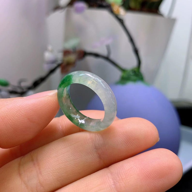 May include: A translucent jade ring with a band of green and white hues. The ring is held between fingers, showcasing its polished surface and natural color variations. The ring is a piece of jewelry.